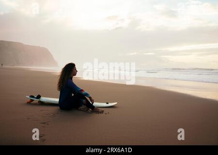 Donna premurosa con tavola da surf seduta in spiaggia Foto Stock