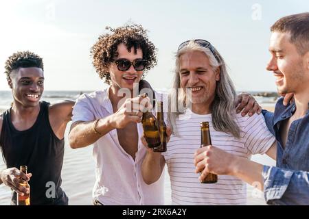 Amici sorridenti che tostano bottiglie di birra in spiaggia nelle giornate di sole Foto Stock