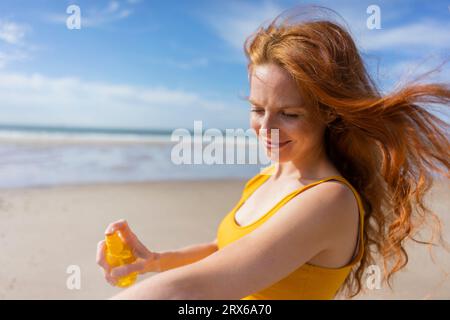 Donna sorridente che spruzza lozione abbronzante a portata di mano in spiaggia Foto Stock