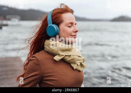 Donna rossa con gli occhi chiusi che ascolta musica tramite cuffie wireless Foto Stock