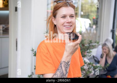 Donna sorridente che invia messaggi vocali tramite telefono cellulare al caffè del marciapiede Foto Stock