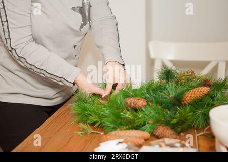 Le mani della donna decorano la corona di natale su un tavolo rustico in legno con coni di pino. Creare una corona di Natale, un'immagine lunatica per le vacanze. Workshop natalizio Foto Stock