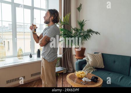 Un giovane premuroso che tiene una bottiglia di birra e guarda dalla finestra Foto Stock