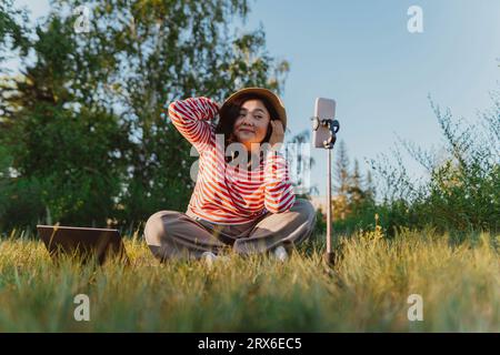 Donna che indossa un cappello e registra vlog sul cellulare sul campo Foto Stock