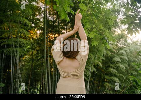 Giovane donna con le braccia alzate in piedi vicino alle piante di bambù Foto Stock