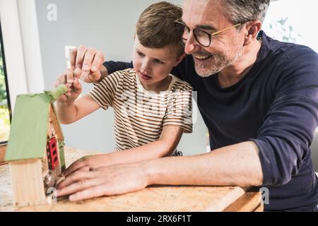 Nonno sorridente che insegna a nipote a dipingere casa modello a casa Foto Stock