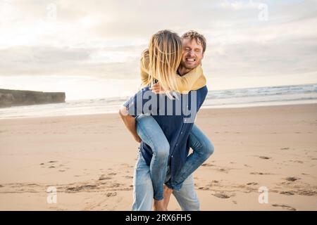 Uomo felice che dà cavalcata piggyback a donna in spiaggia Foto Stock