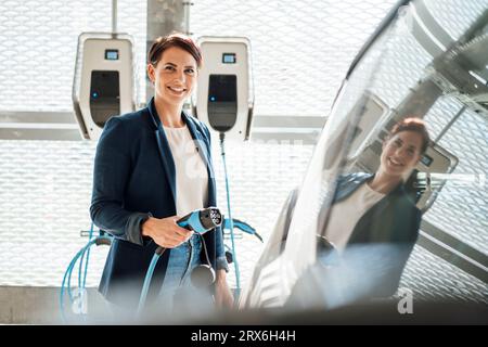 Donna d'affari sorridente con presa elettrica in auto alla stazione di ricarica Foto Stock