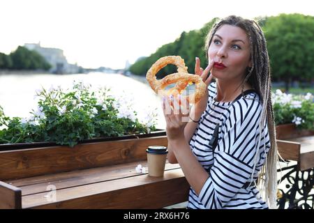 Una giovane donna con un'acconciatura elegante tiene in mano un bagel e le porta un bagel all'occhio con un'espressione giocosa sul viso. Primo piano Foto Stock