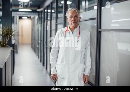 Medico anziano con stetoscopio appoggiato su una parete di vetro in clinica Foto Stock