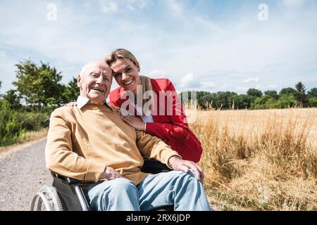 Donna sorridente con un uomo anziano seduto in sedia a rotelle nelle giornate di sole Foto Stock