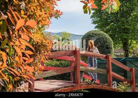 Giardiniera zenzero che cammina su un ponte di legno rosso con rastrello sulla spalla Foto Stock