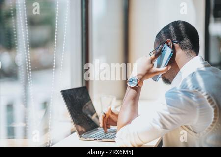 Uomo di colore, di sesso maschile, che ha telefonato mentre lavora sul computer portatile seduto alla scrivania accanto alla finestra dell'ufficio in una giornata di sole. Multi Foto Stock