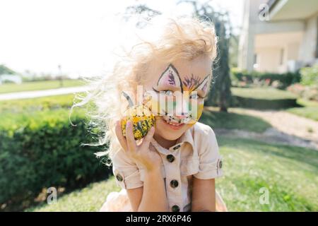 Ragazza felice con la pittura del viso che tiene in mano una piccola zucca nelle giornate di sole Foto Stock