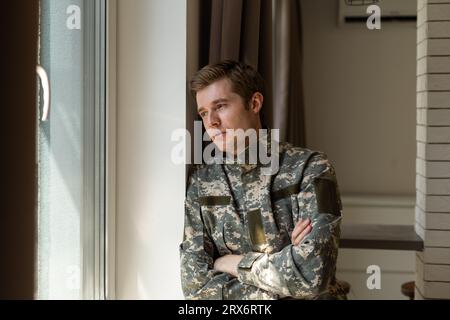 Soldato depresso e triste in uniforme verde con trauma dopo la guerra in piedi vicino alla finestra Foto Stock