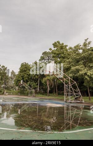 Il tabellone da basket in legno con il cerchio in metallo e la rete bianca mostrano il loro riflesso sull'acqua piovana su tutto il vecchio cemento da basket Foto Stock