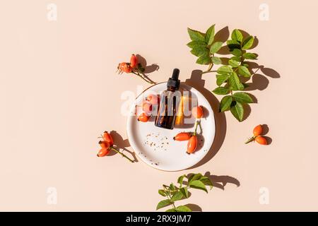 Una bottiglia cosmetica con una pipetta con olio naturale di rosa si trova su un piatto tra le bacche. Vista dall'alto. Uno spazio per le copie. Un concetto naturale di cura di sé Foto Stock