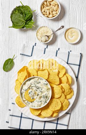 salsa cremosa di spinaci di carciofi e mandorle con patatine fritte su piatto bianco su tavola di legno bianco con ingredienti, vista verticale, posa piatta Foto Stock