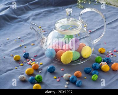 Composizione con uova di pasqua in teiera di vetro circondata da piccole uova di cioccolato e spruzzi dolci. Tè pasquale. Teiera piena di uova colorate Foto Stock