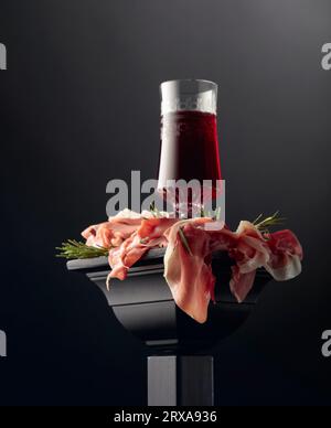 Bicchiere di vino rosso e prosciutto con rosmarino su un podio nero. Foto Stock