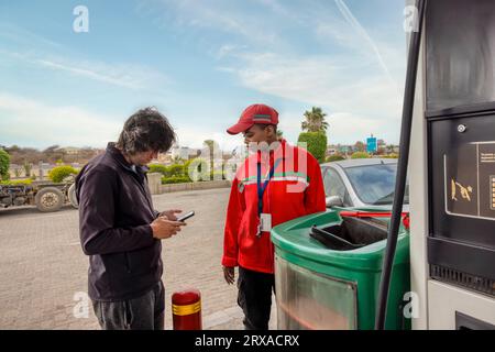 assistente donna afroamericana in uniforme rossa e faccia sorridente alla pompa del gas, cliente che effettua un pagamento mobile con lo smartphone . Foto Stock