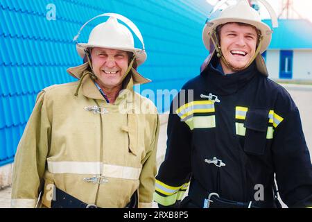 Due pompieri allegri. Due vigili del fuoco in caschi e indumenti protettivi si trovano all'esterno sulla strada e ridono allegramente. Ritratto di felici soccorritori. Foto Stock