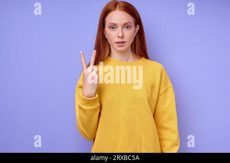 Bella donna seria sicura di sé rossa in elegante maglione giallo che mostra il simbolo della lettera V del linguaggio dei segni per gli umani sordi con sfondo blu. io Foto Stock