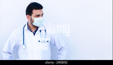 Giovane maschio che indossa maschera e cappotto medico mentre tiene stetoscopio sul collo in modo sicuro Foto Stock