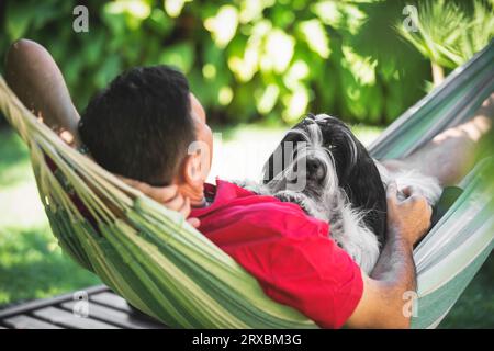 Cane e uomo terrier tibetani giacciono insieme in un'amaca. L'uomo e il suo bellissimo cane che si stendono su un'amaca in campagna, si rilassano durante il giorno Foto Stock