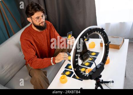 Astrologia online. L'indovinello maschile utilizza uno smartphone e un illuminatore anulare, legge i tarocchi per le previsioni, raccontando ai clienti fornendo servizi Foto Stock