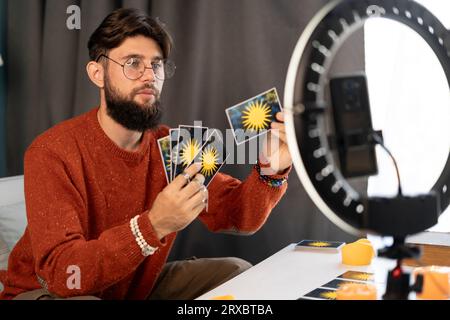 Astrologia online. L'indovinello maschile utilizza uno smartphone e un illuminatore anulare, legge i tarocchi per le previsioni, raccontando ai clienti fornendo servizi Foto Stock