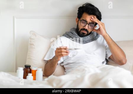 Uomo indiano malato che indossa sciarpa seduto nel letto e tiene in mano il termometro Foto Stock