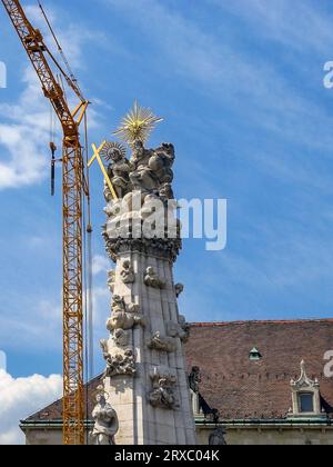 Gru da costruzione vicino alla colonna della Santissima Trinità (colonna della peste) a Budapest contro il cielo blu con le nuvole. La colonna della peste si trova sulla collina del castello in mezzo Foto Stock