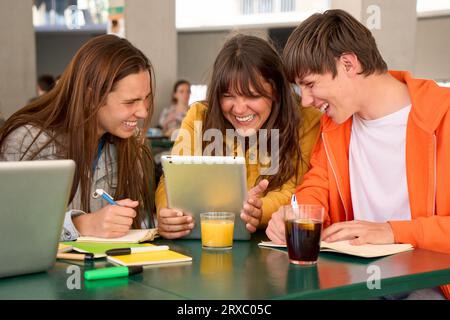 Tre studenti caucasici sorridenti che bevono e fanno i compiti usando il tablet all'università. Foto Stock