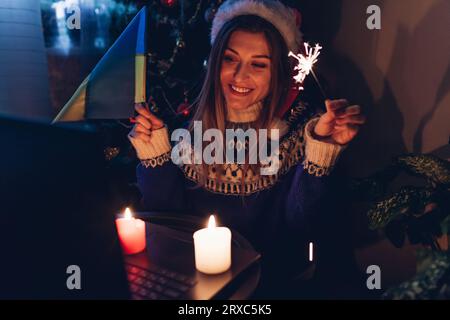 Festeggiamo il Natale e il nuovo anno a distanza online utilizzando il notebook durante la guerra. Donna brucia scintillio a casa tenendo bandiera Ucraina. Che chiama marito Foto Stock