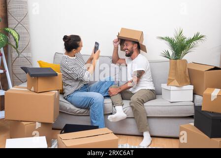 divertente coppia nel giorno in cui ci si muove, disimballare gli effetti personali, divertirsi a scattare foto selfie sul divano in una nuova casa, Foto Stock