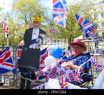 Una signora siede accanto a un cartone tagliato fuori da re Carlo, circondata da bandiere Union Jack, avendo messo al sicuro il suo posto sul Mall di Londra prima dell'evento Foto Stock