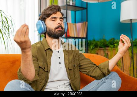 Mantenete la calma, rilassatevi. L'uomo arabo medio-orientale ascoltando la musica respira profondamente, gli occhi chiusi meditano con pensieri concentrati, mente pacifica. Un giovane ragazzo barbuto siede nel salotto di casa sul divano Foto Stock