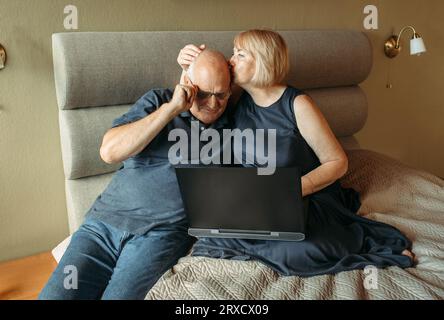 Una felice coppia di pensionati anziani che fanno acquisti online scegliendo insieme gli articoli usando un notebook. L'uomo regola i suoi occhiali. Foto Stock