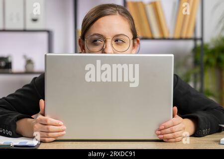 Donna d'affari sicura che si nasconde dietro un computer portatile, guarda la fotocamera, spiando i colleghi che lavorano, piangendo. Ragazza freelance professionista che guarda da dietro il computer con astuto sguardo occhi Foto Stock