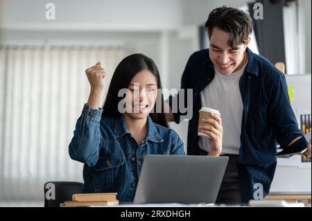 Due giovani imprenditori asiatici festeggiano insieme il successo del loro progetto, urlando di gioia dopo aver ricevuto un'e-mail inaspettata, worki Foto Stock