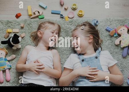 Vista dall'alto dei bambini che giocano con giocattoli colorati. Ritratto di bambini sdraiati sul tappeto nella camera da letto o nell'asilo. Due sorridenti sorridenti Foto Stock