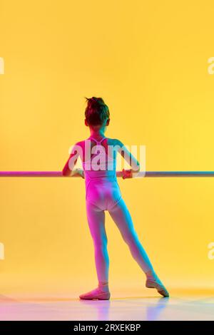 Ritratto retrovisivo di una piccola adorabile ballerina in costume da ballo, legging e pointe che si pratica per lo spettacolo in una scuola di danza con luce al neon Foto Stock