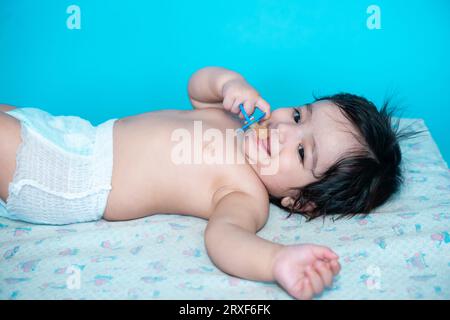 Piccolo bambino indiano di sei mesi che indossa un pannolino sdraiato sul pavimento con capezzolo, succhietto o succhietto isolato sullo sfondo blu dello studio, copia spa Foto Stock