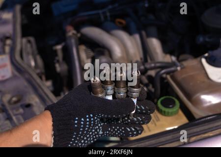 Primo piano di una mano in un guanto nero con quattro candele usate per auto sullo sfondo del motore. Concetto di riparazione auto Foto Stock