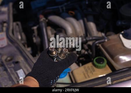 Primo piano di una mano in un guanto nero con quattro candele usate per auto sullo sfondo del motore. Concetto di riparazione auto Foto Stock