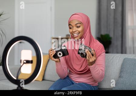 Felice donna islamica afro-americana che fa una recensione su fotocamere retrò, registra video per blog, seduto a casa Foto Stock