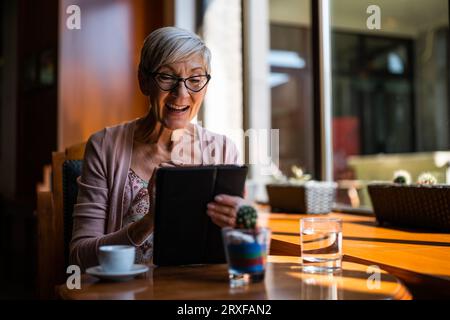 Una donna matura è seduta in una caffetteria e si sta rilassando. Sta usando un tablet digitale. Foto Stock