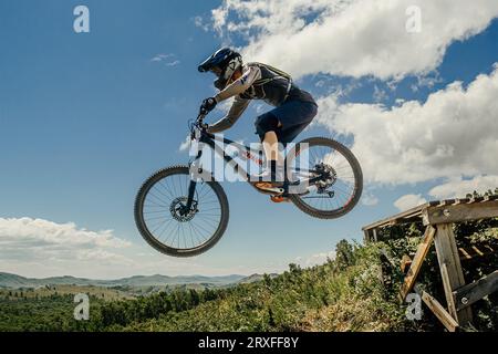 atleta mountain biker saltare giù giù in volo. sullo sfondo del cielo azzurro e delle montagne Foto Stock