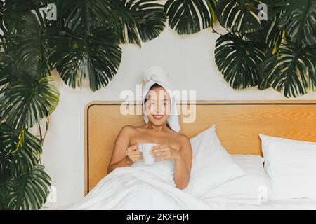 Donna sdraiata a letto con una pianta verde che tiene la tazza del caffè con un asciugamano sulla testa. Foto Stock